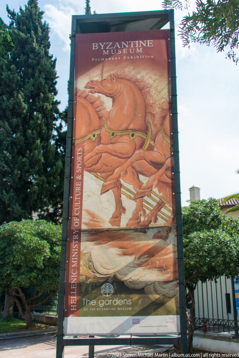 Tall Byzantine Museum banner with a mounted figure reproduction beside hedges at the garden entrance.