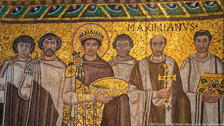 Close view of haloed Emperor Justinian holding a golden vessel among clergy and attendants in a mosaic.