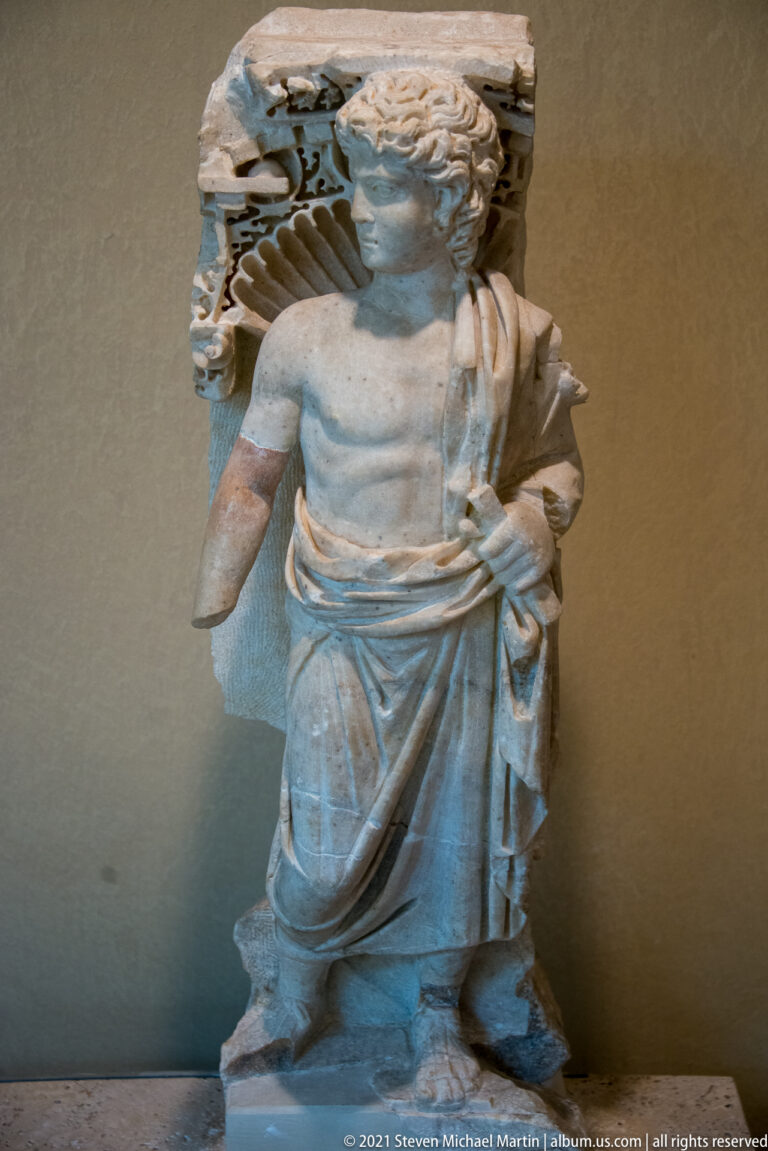 Standing marble figure in draped clothing with a missing arm and carved architectural support behind the head.