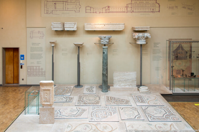 Gallery with columns, capitals, carved stone fragments, patterned mosaic pavement, diagrams, and glass display cases.