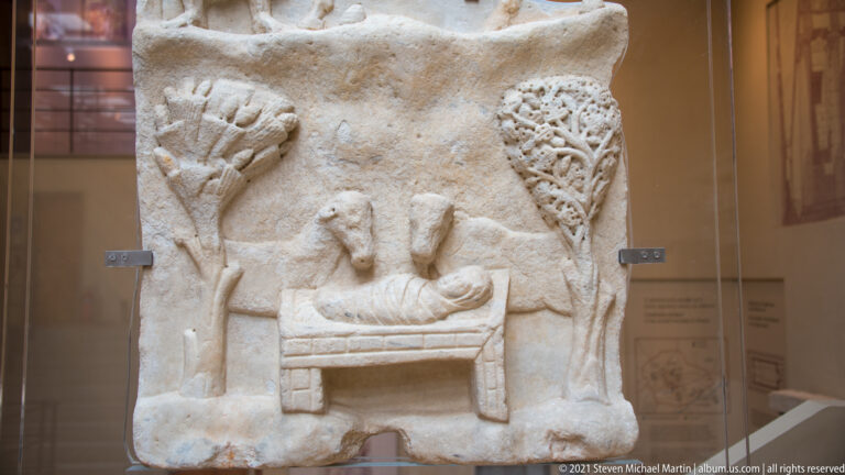 Weathered stone relief with a reclining figure on a couch, smaller figures, and carved motifs around the scene.