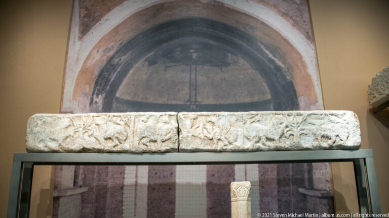 Long carved marble block with repeating vegetal and cross-like motifs displayed before an arched reconstruction.