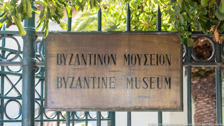 Bilingual Byzantine Museum sign mounted on a decorative metal gate beneath leafy branches.