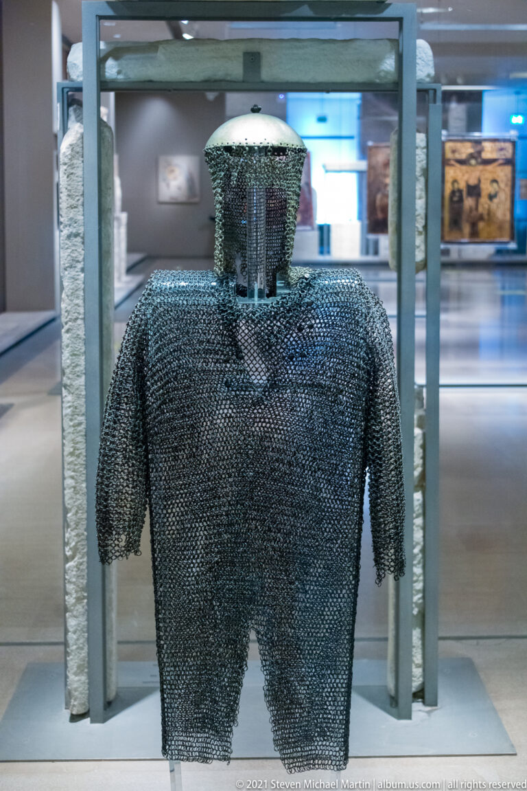 Chain-mail shirt with short sleeves and hood displayed upright inside a museum glass case.