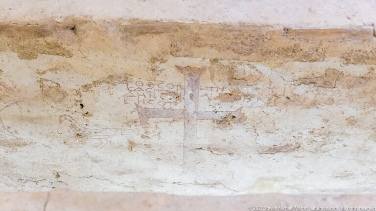 Faint reddish Greek letters and marks on a worn pale plaster or masonry wall surface.