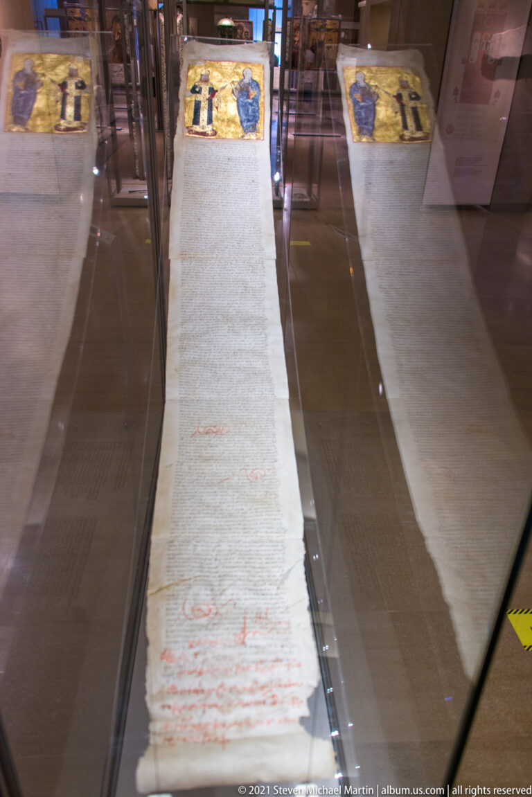 Long manuscript roll in a glass case with red writing and a small painted figural panel at the top.
