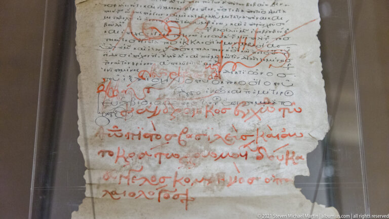 Close-up of a worn Greek manuscript leaf with dense dark writing and prominent red annotations.