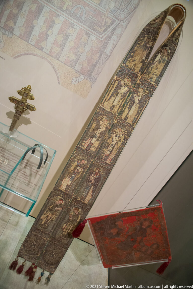 Long narrow painted wooden church furnishing with multiple sacred scenes, displayed horizontally above other museum objects.
