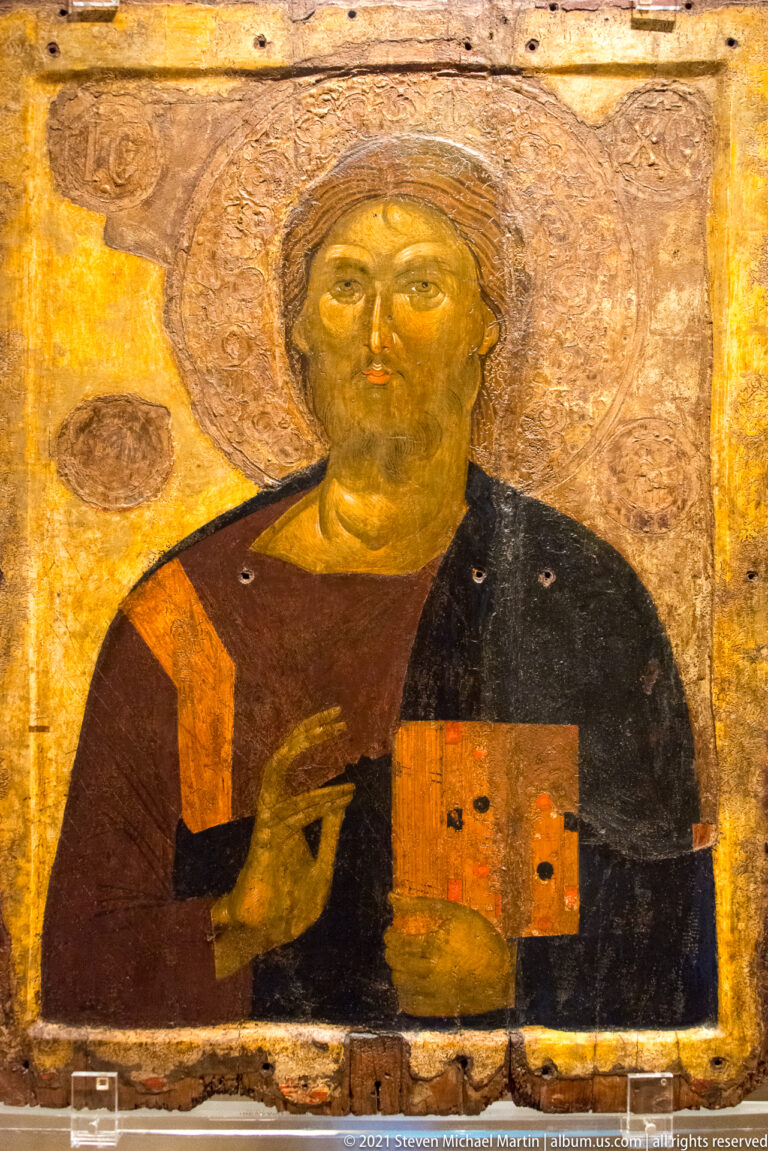 Painted icon of a haloed male saint holding a decorated book and raising his right hand against a gold ground.