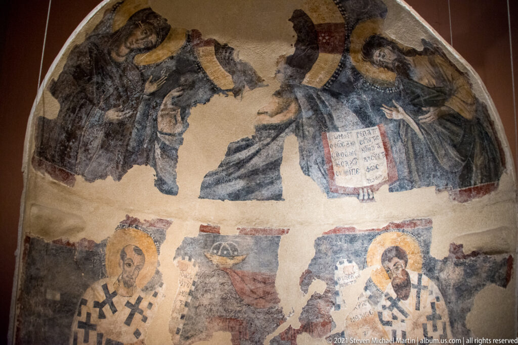 Fragmentary wall painting with haloed clergy in a lower register and a standing figure holding an inscribed text above.