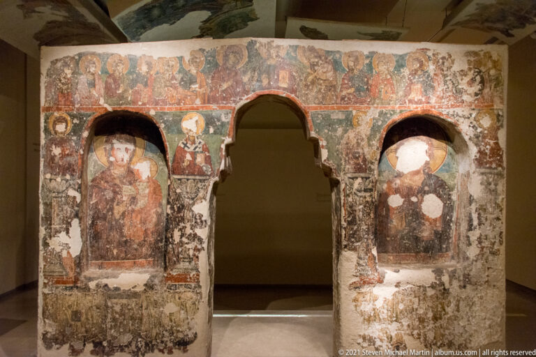 Frescoed masonry fragment with three arched openings, standing saints in side recesses, and a painted figural band above.