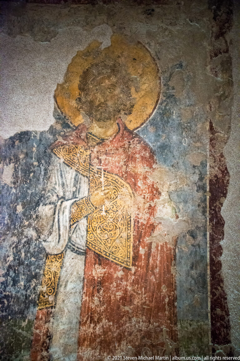 Fragmentary wall painting of a haloed saint in red holding an ornate book, with substantial losses across the face and plaster.