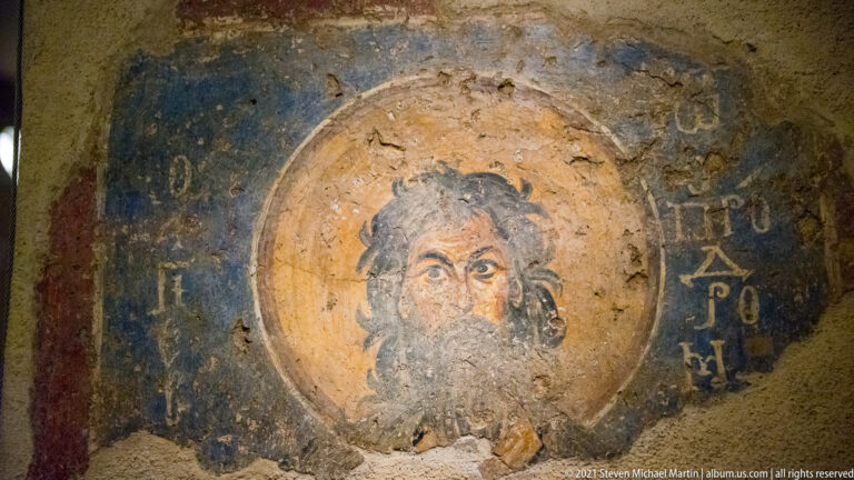 Close view of a bearded haloed saint on blue ground with Greek lettering and extensive cracks and plaster loss.