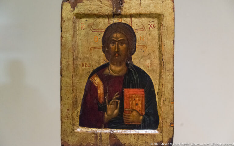 Narrow icon of Christ Pantocrator blessing with his right hand and holding a red book against a gold ground.