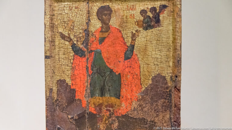Standing female saint in a red mantle with raised hands, small sacred figures above, and extensive cracking across the icon.