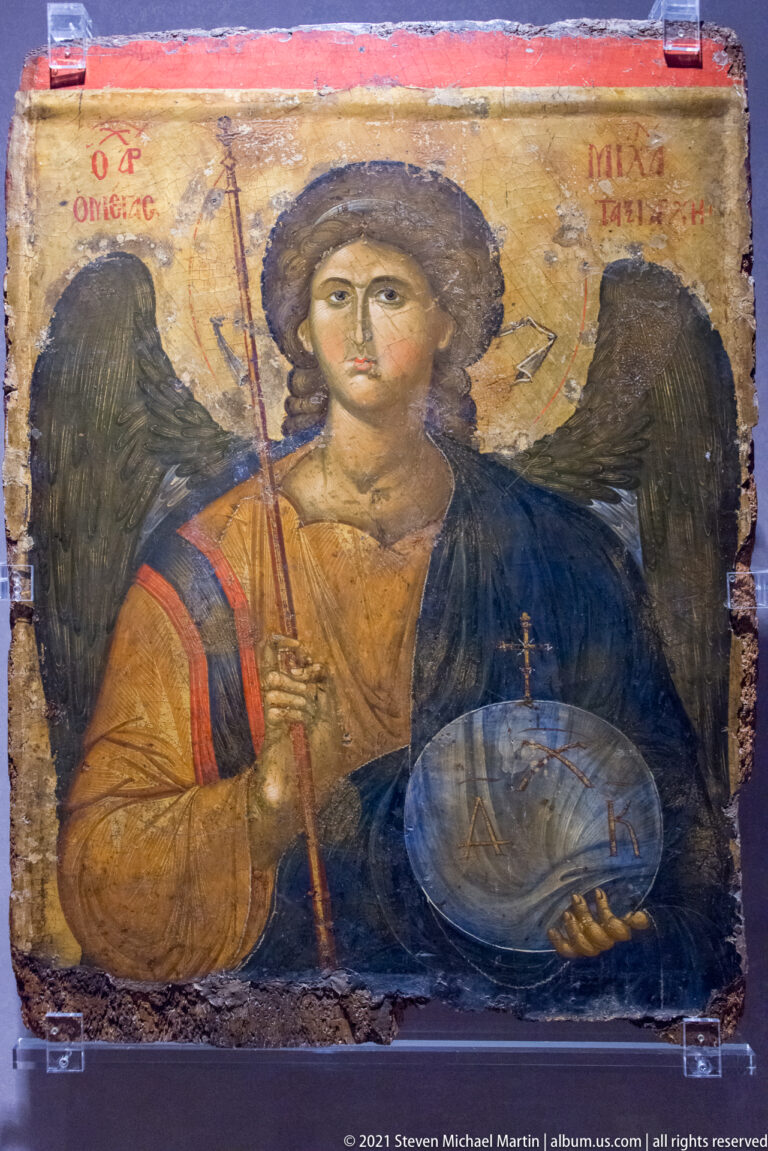 Winged archangel holding a staff and transparent orb, wearing red and blue garments against a worn gold ground.