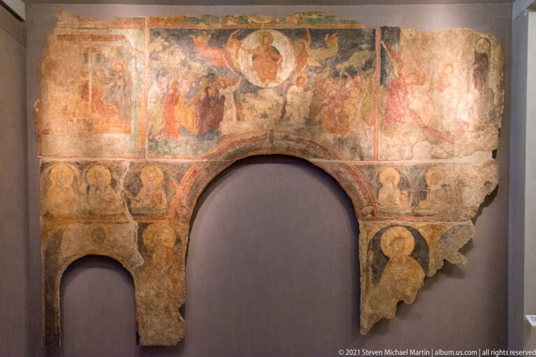 Large frescoed wall fragment with two arched openings, haloed figures, ornamental bands, and irregular areas of missing plaster.