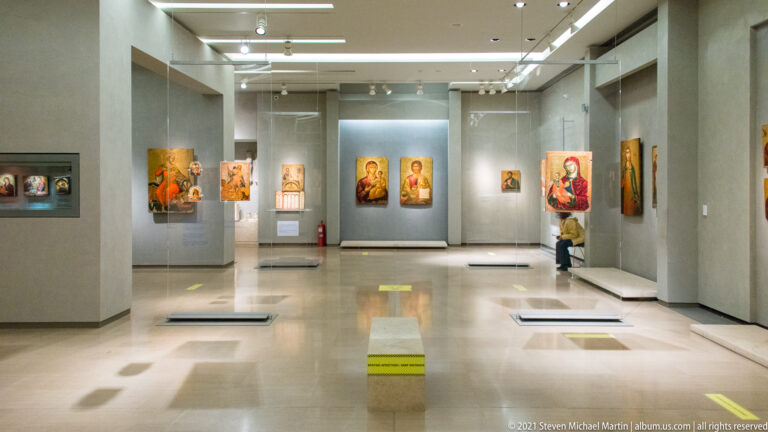 Modern museum gallery with Byzantine icons mounted on gray partitions under ceiling lights, extending through several open exhibition spaces.