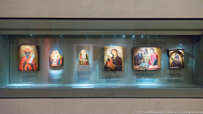 Illuminated museum case displaying six small religious icons of saints and multi-figure scenes with labels beneath.