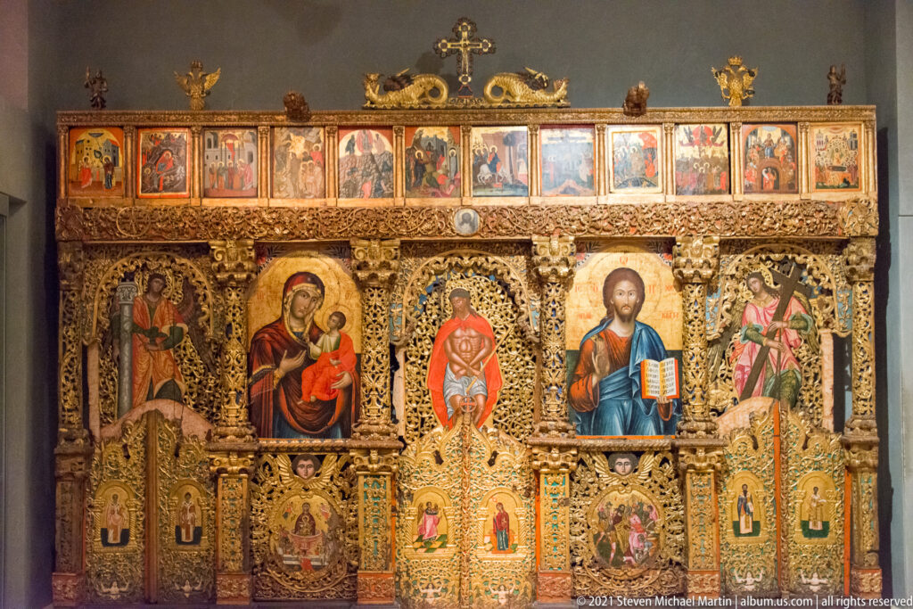 Large gilded carved iconostasis with major icons, a row of smaller narrative panels, ornamental columns, and a cross at top.