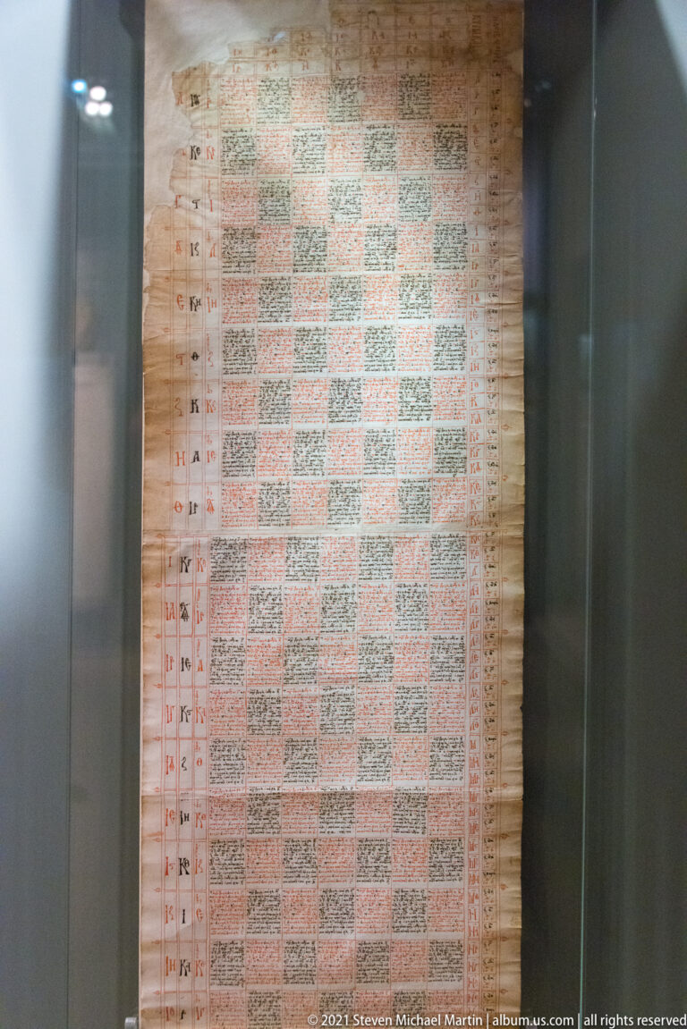 Tall manuscript or printed sheet with a dense grid of pale and red text-filled cells, displayed vertically behind glass.