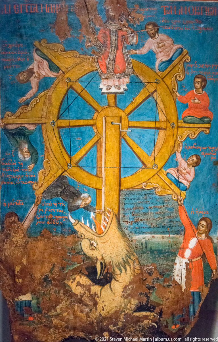 Colorful manuscript illustration of a large golden wheel with figures around it, an animal below, and red handwritten labels on blue ground.