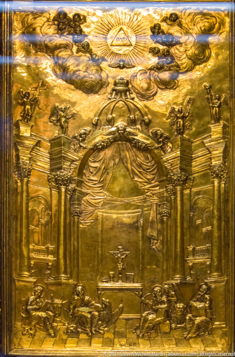 Gilt-metal relief showing an ornate columned sacred interior beneath clouds and a radiant triangle, with small figures and furnishings below.