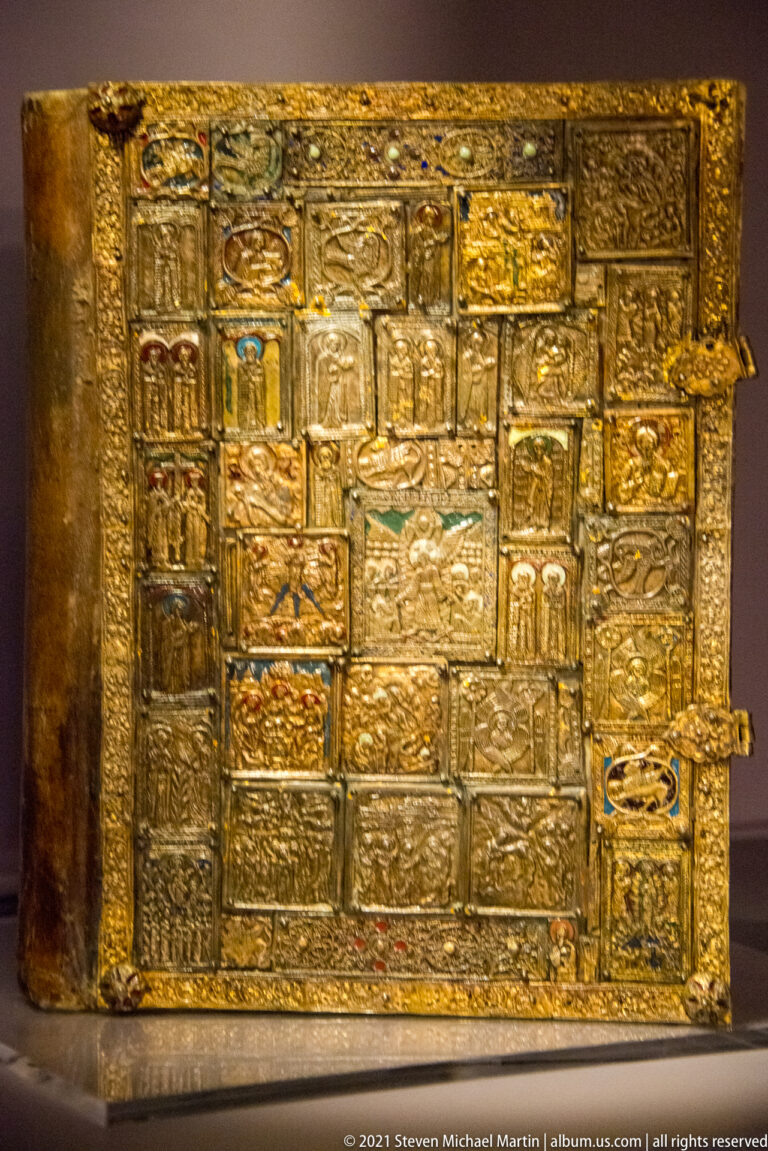 Rectangular gilt-metal book binding with many embossed narrative panels, ornamental borders, hinges, and a projecting clasp at right.