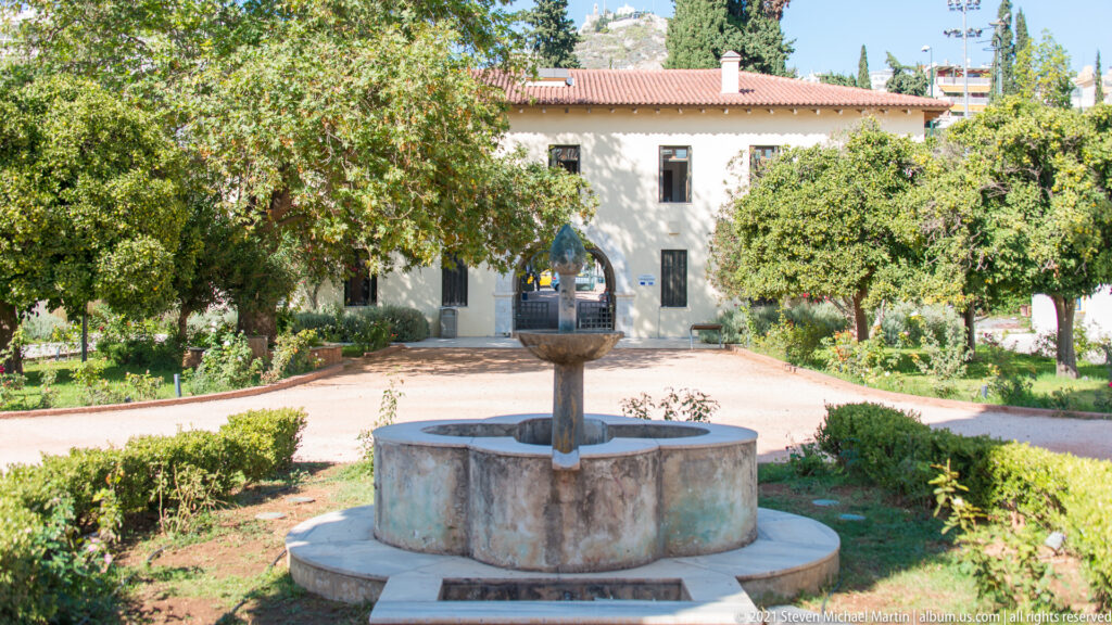 Circular stone fountain centered in a garden courtyard before a pale, red-tiled museum building with an arched entrance.