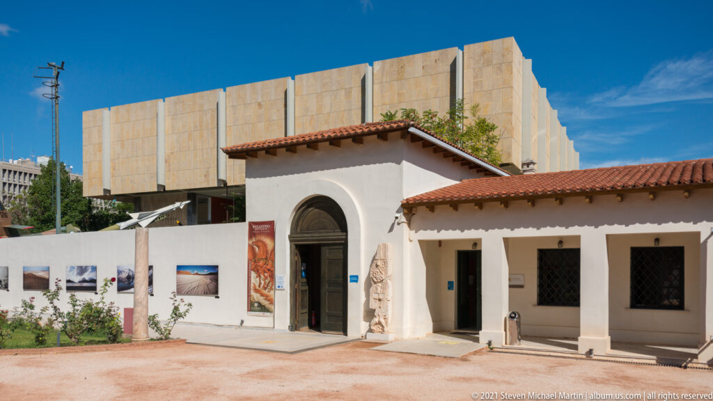 White museum entrance pavilion with arched doorway and red tile roof in front of a large modern building.