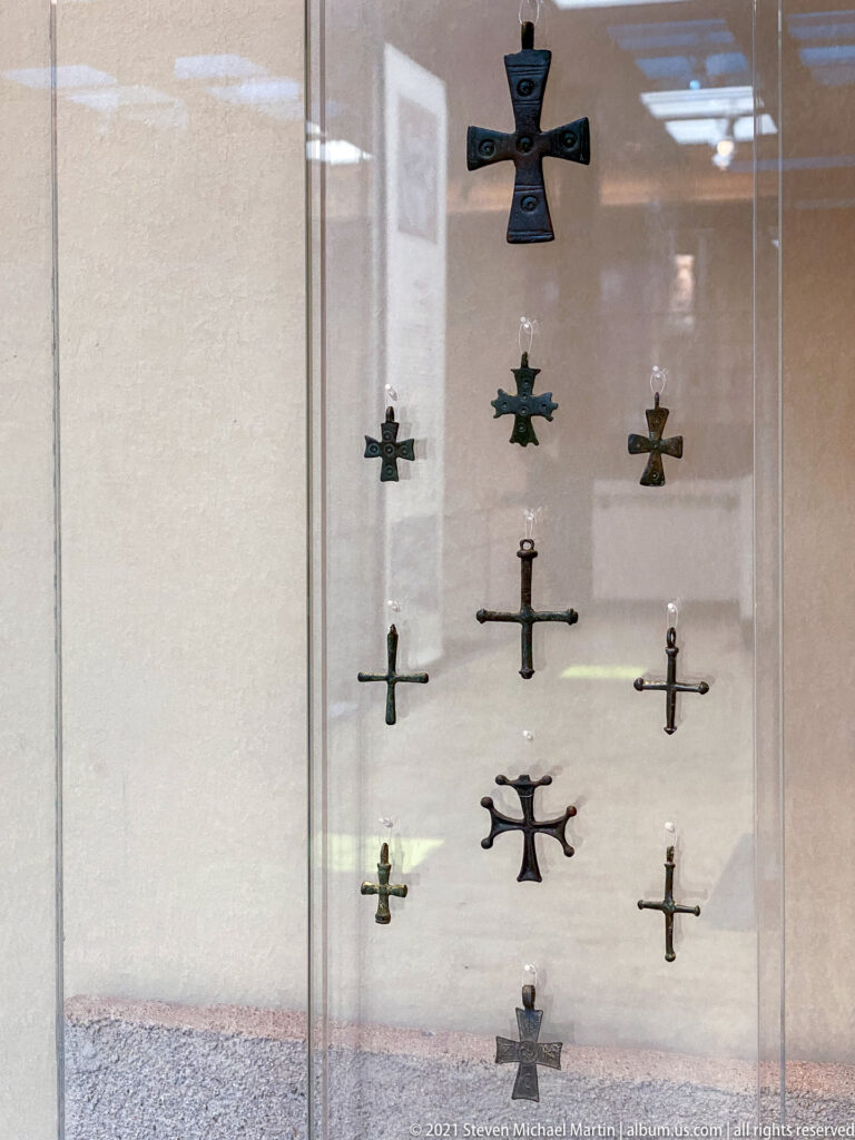 Glass case displaying many small metal crosses of different shapes arranged in vertical rows.