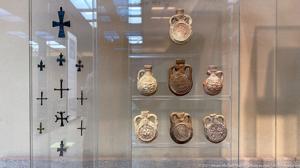 Small rounded ceramic flasks with relief medallions displayed on shelves beside a case of metal crosses.