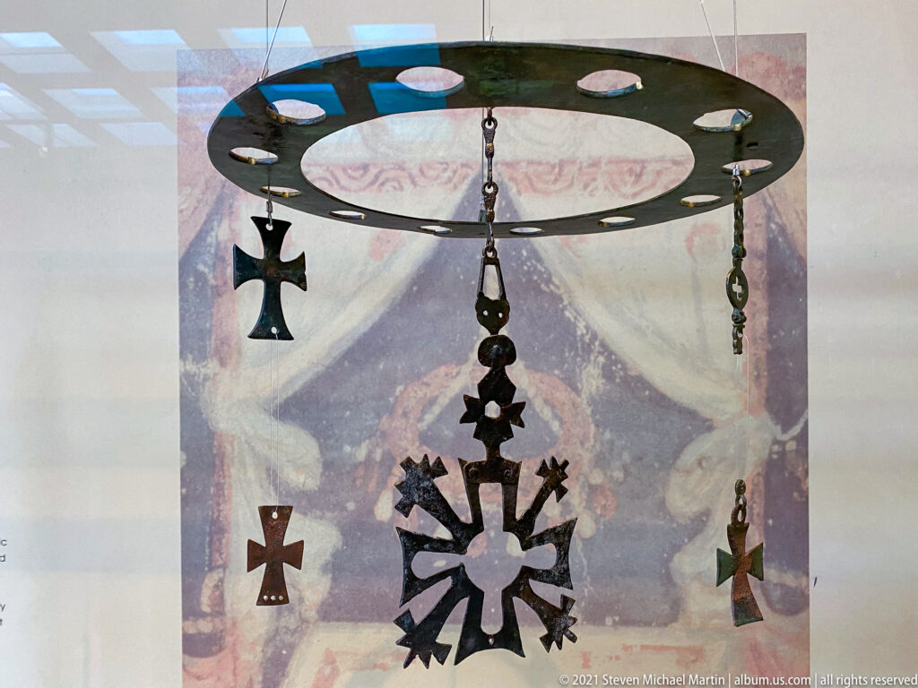 Circular suspended metal fitting with chains and multiple cross-shaped pendants hanging before a painted background.