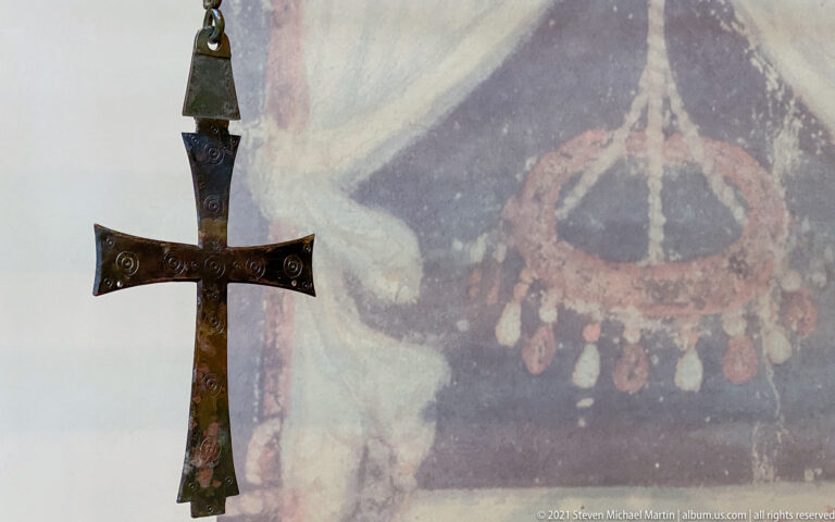 Dark metal pendant cross with engraved circular ornament suspended in front of a faded wall painting.