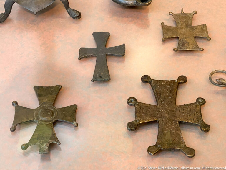 Four small cast metal crosses with flared and rounded arm ends displayed flat in a case.