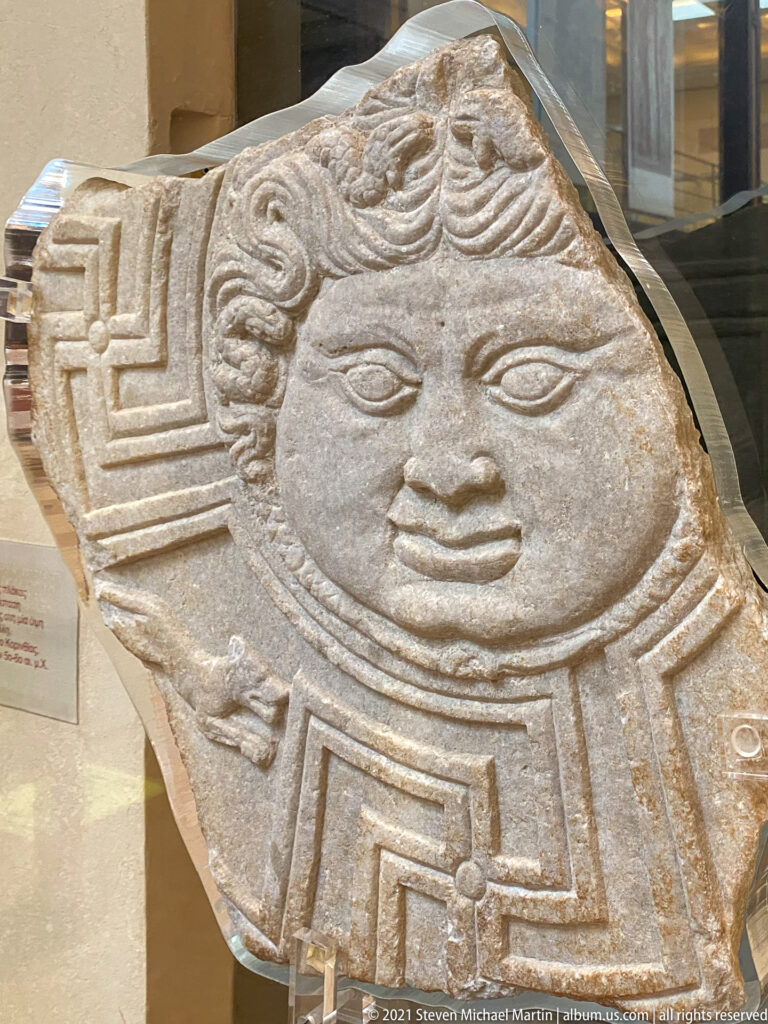 Stone relief fragment with a large frontal human face surrounded by angular geometric carving.