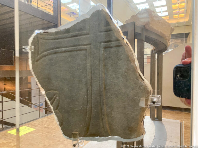 Upright broken stone fragment with a deeply recessed cross and stepped linear carving, displayed on a stand in the museum.