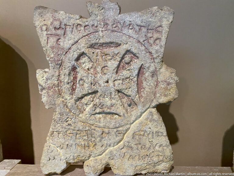 Irregular stone fragment with a central cross motif, faint lettering, and traces of reddish pigment.