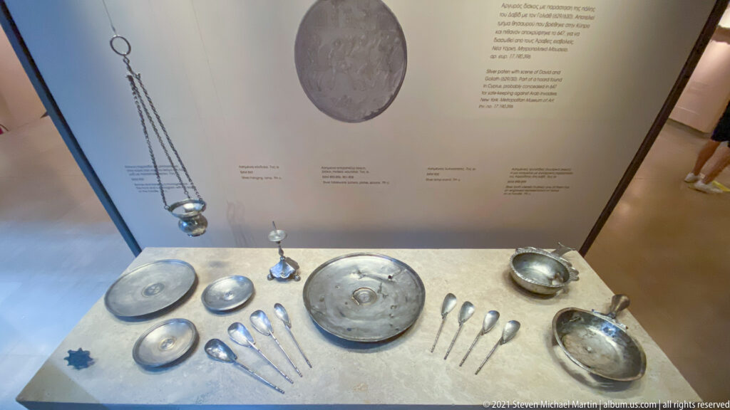 Museum case with silver-colored plates, bowls, spoons, small vessels, and a long chain arranged beneath labels.