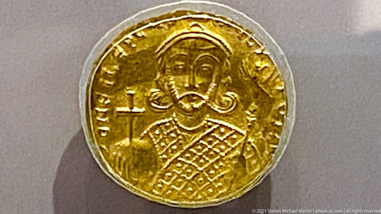 Close-up of a gold coin bearing a frontal ruler in patterned dress beside a cross and encircling inscription.