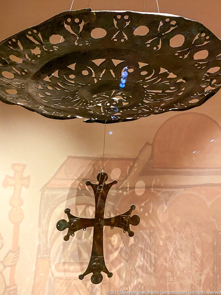 Large perforated metal disk suspended horizontally above a hanging cross, casting a shadow on the museum wall.