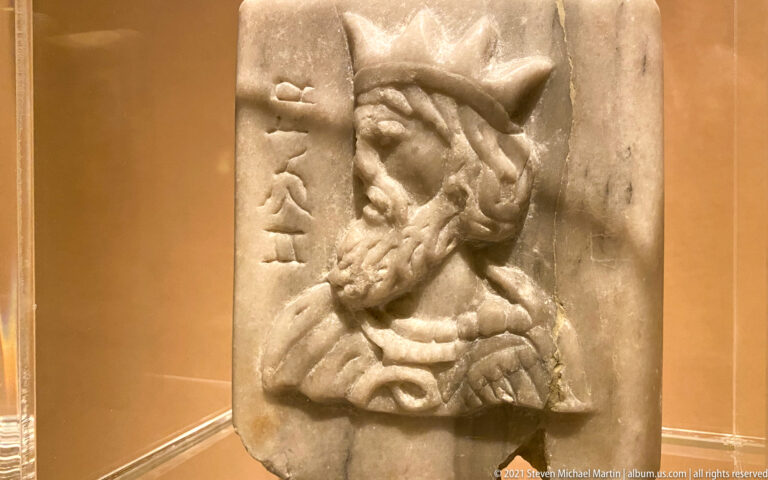 Small marble relief bust of a bearded man in a pointed head covering beside a vertical inscription.