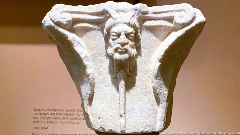 Pale marble architectural block with broad projecting forms framing a deeply carved bearded male head.