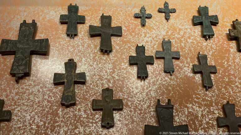 Group of dark metal pendant crosses of many sizes and shapes mounted against a mottled museum display surface.