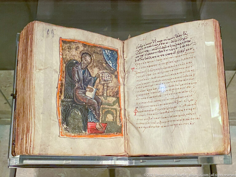 Open Greek manuscript with a painted haloed seated writer on the left and dense handwritten text on the right.