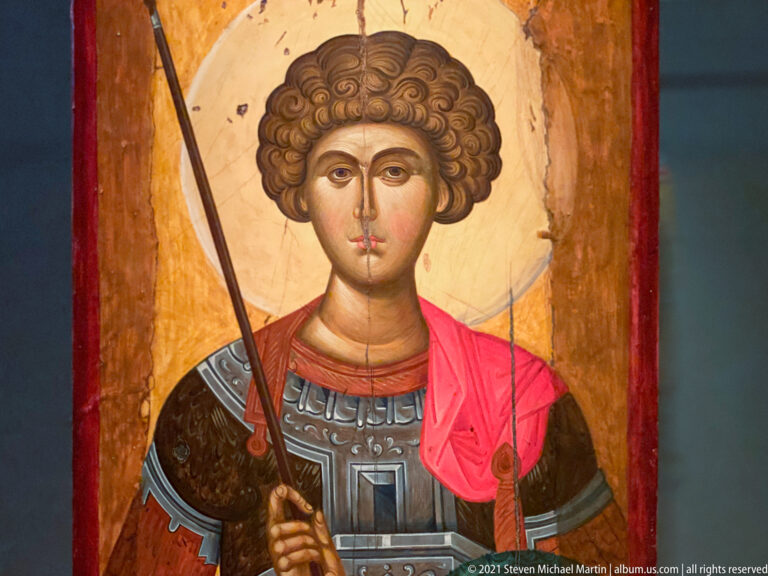 Close view of a Byzantine icon showing a youthful warrior saint with curled hair, spear, shield, armor, and red cloak.