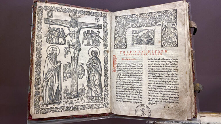 Open Greek religious book with two-column text, red headings, full-length sacred figures, narrative scenes, and ornate illustrated borders.