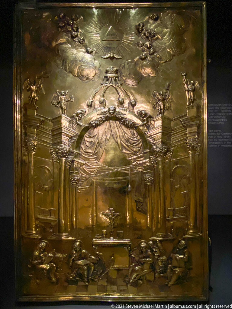 Large gilded metal ecclesiastical object covered with repoussé figures, arches, scrolling ornament, and architectural details in a museum case.
