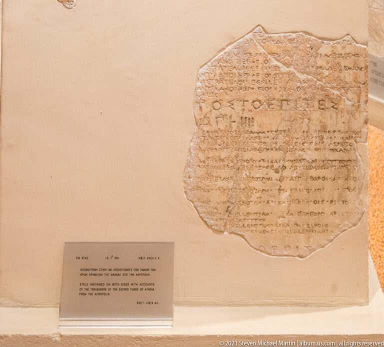 Irregular pale stone fragment covered with many horizontal lines of ancient Greek lettering in a museum case.