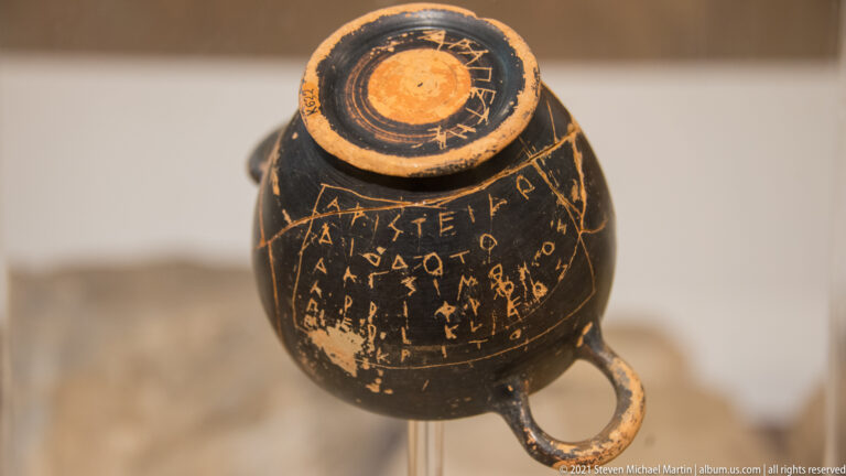 Small black ceramic vessel with a handle and several lines of scratched Greek letters across its body.